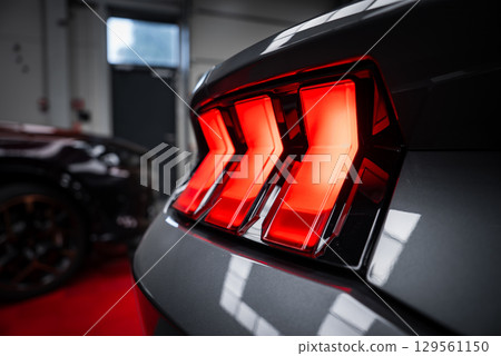 Close up of the illuminated tri bar LED taillight of a Ford Mustang GT 5.0. The glossy black exterior reflects showroom lighting, with another car in the background. Close up of the illuminated tri bar LED taillight of a Ford Mustang GT 5.0. The glossy black exterior reflects showroom lighting, with another car in the background. 129561150