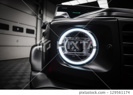 Close up view of a Mercedes AMG G Class headlight with circular LED light, matte black finish, and reflections in a tiled indoor showroom. 129561174