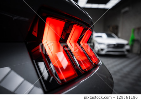 Rear taillight of Ford Mustang GT 5.0 with tri bar LED design illuminated. A white Mercedes AMG G Class is visible in a modern showroom setting. 129561186