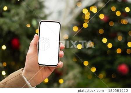 Man hand with smartphone with white blank screen against Christmas tree light garland and fairy lights. Festive Xmas Background. Mock up. Close up. 129561620