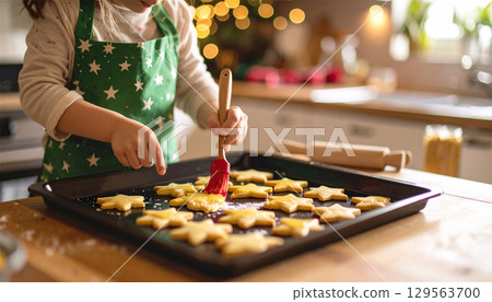 一位戴著綠色頭巾、穿著星形圍裙的小麵包師傅正在刷星形餅乾。 一位戴著綠色頭巾、穿著星形圍裙的小麵包師傅正在刷星形餅乾。 129563700