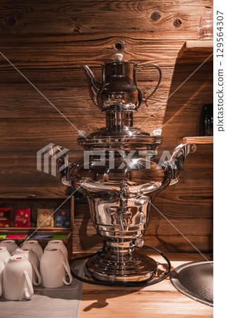 Polished samovar with teapot on top, surrounded by white mugs and tea packets, set against a wooden wall under warm lighting in a cozy environment. Polished samovar with teapot on top, surrounded by white mugs and tea packets, set against a wooden wall under warm lighting in a cozy environment. 129564307