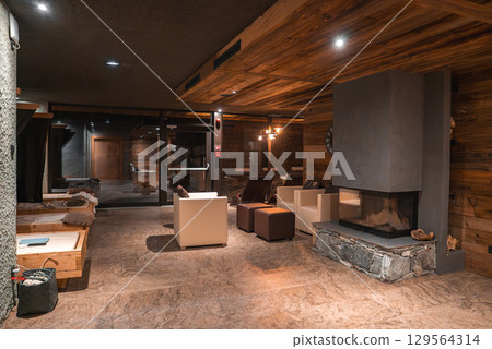 Modern rustic room with wooden walls, stone fireplace, white armchairs, dark ottomans, polished stone flooring, and large glass doors in the Italian Alps. 129564314