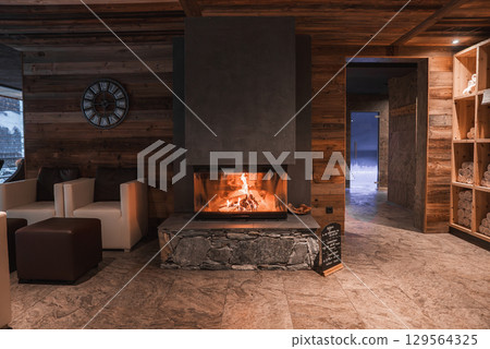 Warm room with wooden walls, modern fireplace, armchairs, and spa shelves. A doorway reveals stone walls and a snowy outdoor view. Warm room with wooden walls, modern fireplace, armchairs, and spa shelves. A doorway reveals stone walls and a snowy outdoor view. 129564325