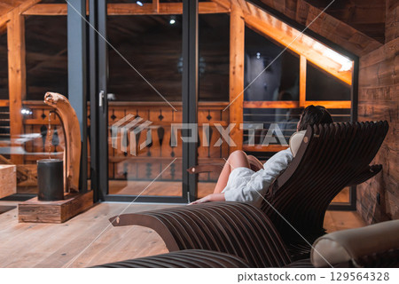 Cozy chalet interior featuring natural wood textures, a person in a white robe on a lounge chair, and glass doors opening to a balcony with wooden railings. 129564328