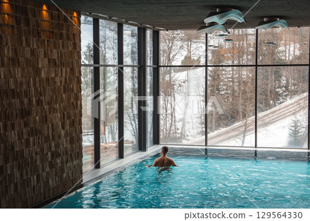 A person swims in an indoor pool with large windows revealing a snowy forest. The space features wooden walls and modern light fixtures, set in the Italian Alps. A person swims in an indoor pool with large windows revealing a snowy forest. The space features wooden walls and modern light fixtures, set in the Italian Alps. 129564330