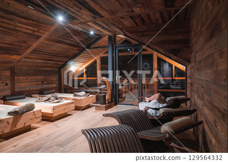 Cozy relaxation room in a luxury hotel in the Italian Alps, featuring sloped wooden ceilings, modern lounge chairs, and large glass doors to a balcony. Cozy relaxation room in a luxury hotel in the Italian Alps, featuring sloped wooden ceilings, modern lounge chairs, and large glass doors to a balcony. 129564332