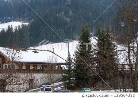 Snowy alpine scene featuring a luxury hotel with stone walls, wooden accents, a ski lift on a forested slope, and parked vehicles in the foreground. 129564338