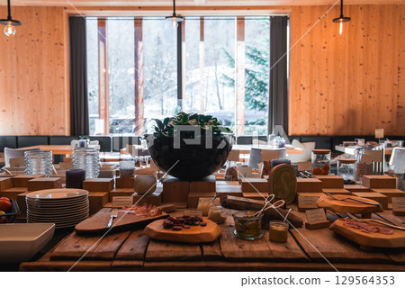 Cozy wooden dining area featuring a buffet with charcuterie, cheeses, and condiments. Large windows reveal snow dusted trees and alpine scenery. 129564353