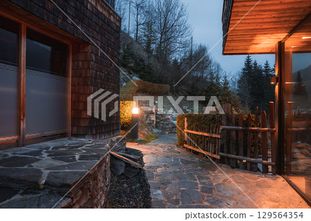 Cozy outdoor space of a luxury hotel in the Italian Alps, featuring a stone pathway, warm lighting, rustic wooden fence, and hilly backdrop with trees. 129564354