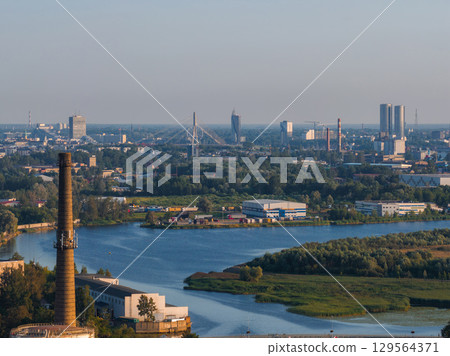 Riga, Latvia, featuring the Daugava River, Vansu Bridge, Swedbank building, industrial chimney, urban skyline, and surrounding greenery in daylight. Riga, Latvia, featuring the Daugava River, Vansu Bridge, Swedbank building, industrial chimney, urban skyline, and surrounding greenery in daylight. 129564371