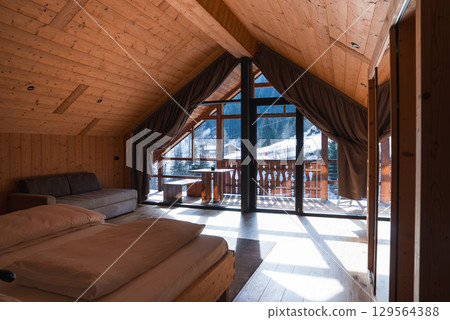 Wooden interior of a hotel room with sloped ceiling, large windows, and balcony overlooking snow covered rooftops and forested mountains. 129564388