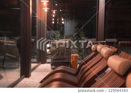 Indoor relaxation area with curved wooden lounge chairs, warm pendant lighting, and large windows overlooking evergreen trees in the Italian Alps. 129564393