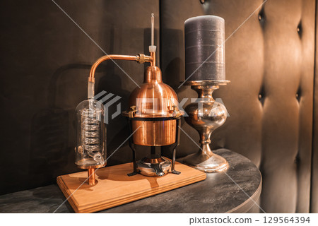 A copper distillation apparatus with glass coil on a wooden base, paired with a dark gray candle in a silver holder against a tufted leather backdrop. 129564394