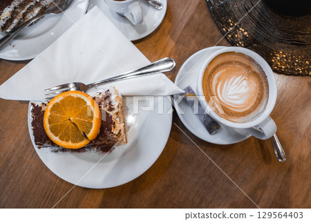 A slice of tiramisu with orange garnish on a white plate, paired with a cappuccino featuring latte art, set on a wooden table in a luxury setting. 129564403