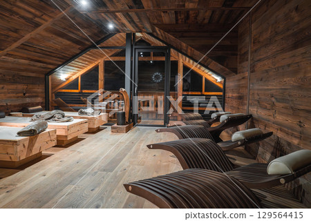Rustic modern room with wooden walls, floors, and ceilings, featuring curved lounge chairs, wooden beds, and a glass door overlooking a balcony at night. 129564415