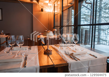 Dining area features wooden tables with white tablecloths, cutlery, and lamps. Large windows reveal a snowy forest, blending modern and rustic design. 129564473