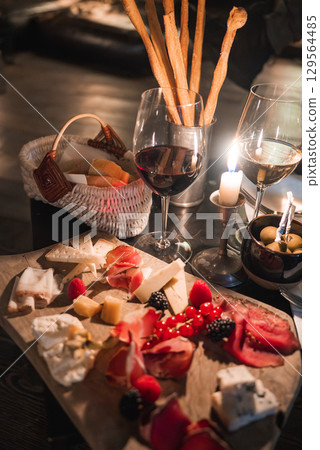 Wooden platter with cheeses, cured meats, berries, and wine on a rustic table. Lit candle and breadsticks add warmth in an Alpine hotel setting. Wooden platter with cheeses, cured meats, berries, and wine on a rustic table. Lit candle and breadsticks add warmth in an Alpine hotel setting. 129564485
