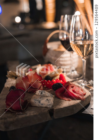 A rustic wooden platter features cured meats, blue cheese, berries, and bread. Red and white wine glasses add elegance in a cozy, dimly lit ambiance. 129564489