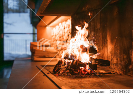 Cozy indoor fireplace with stacked firewood, warm lighting, and a large window showcasing a snowy winter scene in the Italian Alps. Cozy indoor fireplace with stacked firewood, warm lighting, and a large window showcasing a snowy winter scene in the Italian Alps. 129564502