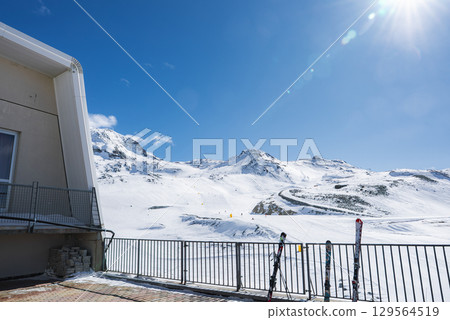 Snow covered slopes at Zermatt ski resort in Switzerland, with a modern building, ski tracks, orange markers, and skis resting on a metal railing. 129564519