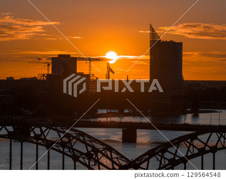 The Riga TV Tower and city skyline stand out at sunset, with the Daugava River and a bridge in the foreground. A Latvian flag is visible near the center. 129564548