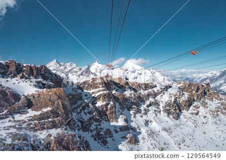 Snowy alpine peaks and rugged terrain with orange gondolas on ski lifts at Zermatt ski resort, set against a clear blue sky and majestic mountains. Snowy alpine peaks and rugged terrain with orange gondolas on ski lifts at Zermatt ski resort, set against a clear blue sky and majestic mountains. 129564549
