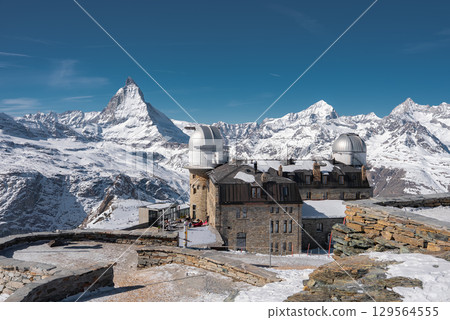The Gornergrat Observatory sits on a rocky plateau in Zermatt, Switzerland, surrounded by snow covered peaks, including the Matterhorn, under a clear blue sky. 129564555