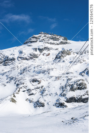Snowy mountain peak in Zermatt, Switzerland, featuring ski lift cables, rocky outcrops, and a summit structure under a bright blue sky. 129564576