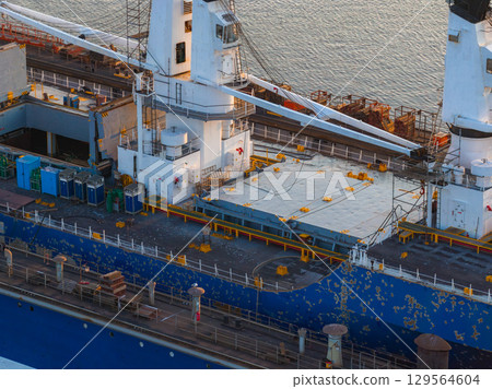 A ship with a blue hull and visible wear docked at a shipyard in Riga, Latvia. Cranes and containers are on the deck, with calm water at sunset. 129564604