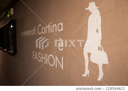 Wall with beige textured finish featuring 'benvenuti Cortina e FASHION' text and a white silhouette of a woman with a hat and handbag. Wall with beige textured finish featuring 'benvenuti Cortina e FASHION' text and a white silhouette of a woman with a hat and handbag. 129564621