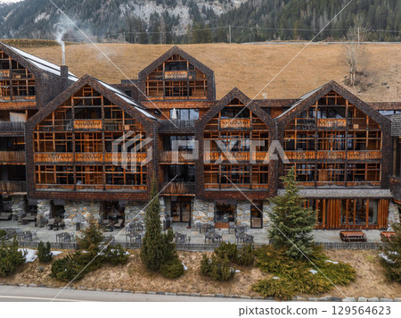 Charming Alpine hotel with rustic wooden design, balconies, and chimney smoke, set amidst evergreen trees, grassy slopes, and dense forest in Italy. 129564623