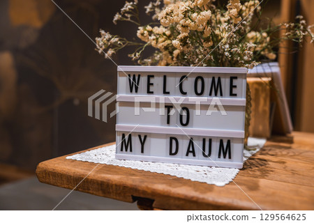 A wooden table features a lightbox sign reading 'WELCOME TO MY DAUM' with a lace doily and dried flowers. Warm tones create a cozy atmosphere. 129564625