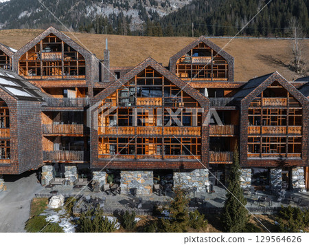 Charming Alpine hotel with steep roofs and wooden balconies, surrounded by forested slopes, rocky peaks, and patches of snow in the Dolomites, Italy. 129564626