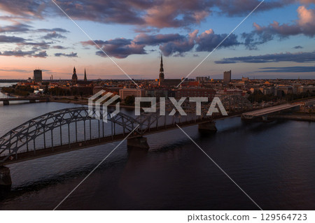 Riga, Latvia at sunset with the Daugava River, Railway Bridge, and spires of Riga Cathedral and St. Peter's Church under a colorful evening sky. 129564723