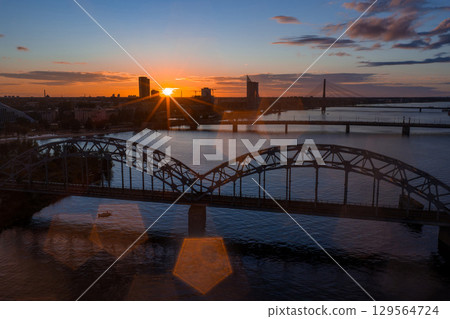 The image shows a sunset over Riga, Latvia, with the Railway Bridge spanning the Daugava River and the Vansu Bridge visible in the background. 129564724