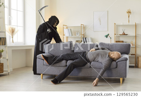Sinister figure of grim reaper of death with scythe standing behind lifeless man on couch. 129565230