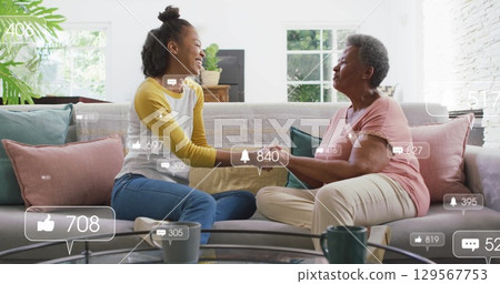 Holding hands mother and daughter sitting on gray sofa in living room, with social media icons 129567753