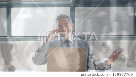 Smiling man in business suit holding smartphone to ear and gesturing at office desk, with laptop 129567769