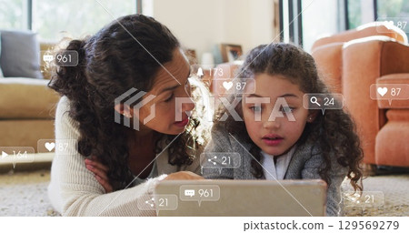 Lying mother and daughter scrolling tablet on living room carpet, with floating social media icons 129569279