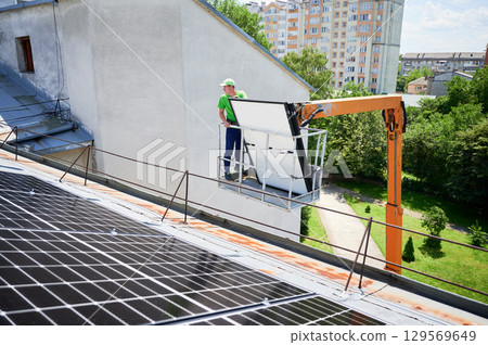 Workers lifting up photovoltaic solar panel on metal rooftop of house with assistance of crane lift. Men installers installing photovoltaic solar modules outdoors. Renewable energy generation concept. Workers lifting up photovoltaic solar panel on metal rooftop of house with assistance of crane lift. Men installers installing photovoltaic solar modules outdoors. Renewable energy generation concept. 129569649