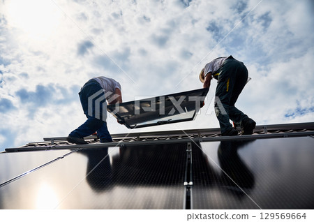 Workers building solar panel system on roof of house. Two men installers in helmets carrying photovoltaic solar module outdoors. Alternative, green and renewable energy generation concept. 129569664