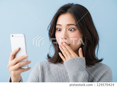 A woman is surprised at her smartphone and covers her mouth with one hand 129570424