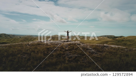 Back view of a young female tourist standing with raised arms on top of a hill and enjoying the beautiful landscape of green hills under a blue cloudy sky Back view of a young female tourist standing with raised arms on top of a hill and enjoying the beautiful landscape of green hills under a blue cloudy sky 129572894