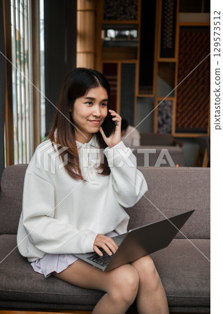 Smiling young woman multitasking with a laptop and smartphone in a stylish cafe with wooden decor. 129573512