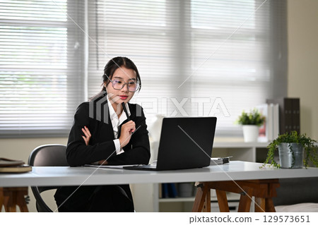 Confident Asian woman in glasses sitting at a desk in office, focused on laptop screen 129573651