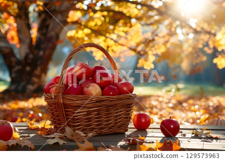 Wicker basket full of red apples sitting on wooden table in autumnal park 129573963