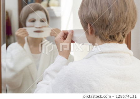 Senior woman taking care of her skin with a face mask in front of the mirror 129574131