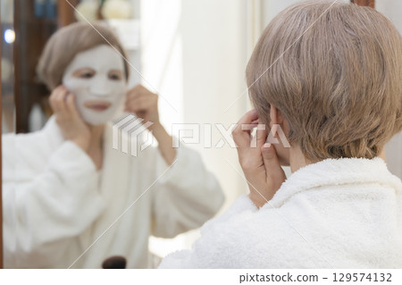 Senior woman taking care of her skin with a face mask in front of the mirror 129574132