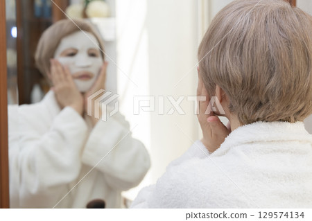 Senior woman taking care of her skin with a face mask in front of the mirror Senior woman taking care of her skin with a face mask in front of the mirror 129574134